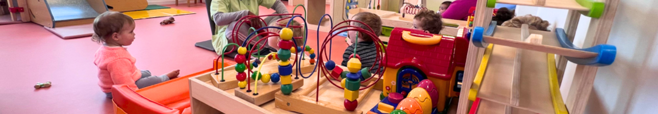 Des bambins assis dans une salle de jeux devant une agent de crèche qui s'adresse à eux, il y a des jouets multicolores en premier plan.