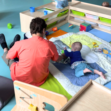 Une agent de crèche avec une blouse rouge assise sur un tapis de jeux pour enfants bleu et vert s'occupe d'un bébé allongé sur le ventre à sa gauche sur le tapis.